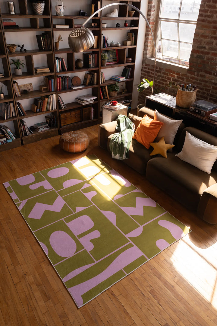 Olive green Abstract Patchwork Rug by Becca Franks in a loft-style room with brick wall, bookshelf, and brown sofa