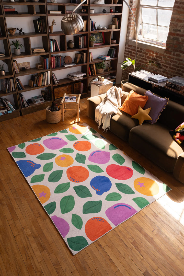 Pastel Fruit Party Rug by Becca Franks laid out in front of a sofa, sunlight casting over its colorful pattern on the wood floor