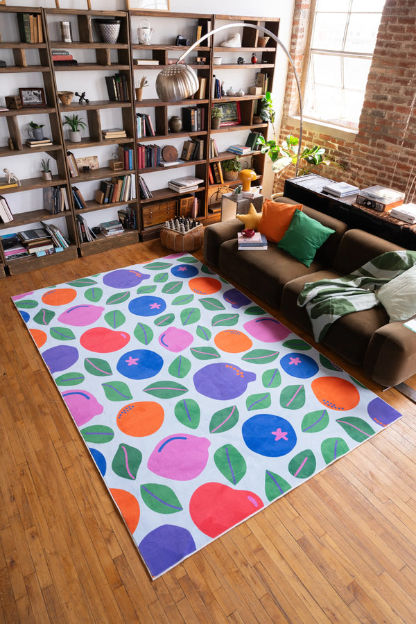 Blue Fruit Party Rug by Becca Franks laid out in a bright room with bookshelves and a brown sofa on wood floor