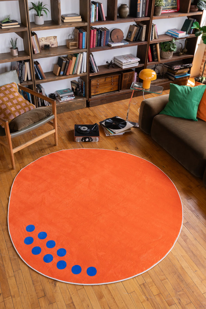 Large orange minimalist rug with blue dot accents centered in a living room with bookshelves and mid-century furniture