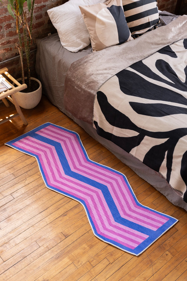 Pink Blue Zigzag Wave Rug by Jakob Mizrahi placed beside bed in cozy bedroom with wood floor