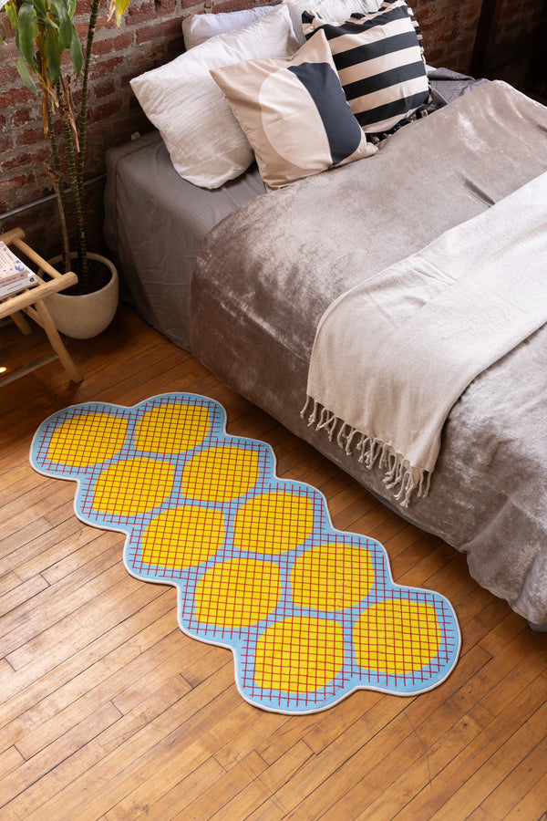 Yellow Grid Lemon Rug by Johann Banta beside neutral bed with throw pillows in cozy brick-walled bedroom