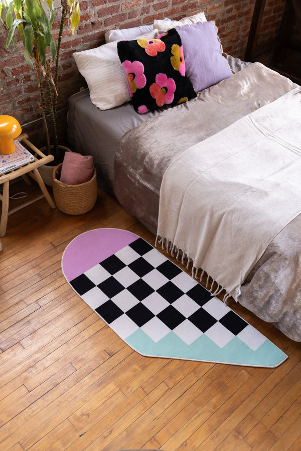 Pink and Blue Asymmetrical Checker Rug by Jakob Mizrahi placed beside a low bed in a bedroom with wood floor and brick wall