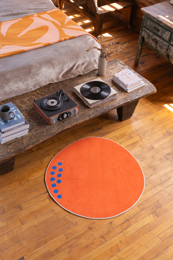 Round orange minimalist rug with blue dot accents placed beside a wooden bed in a vintage-themed bedroom with record player