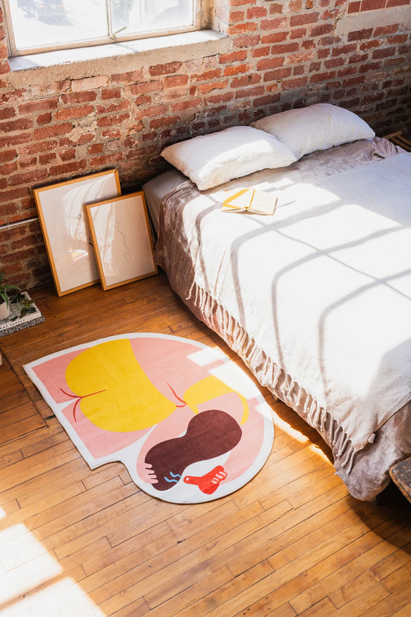 Round body art rug in bold yellow and pink tones catching the sunlight, placed at the foot of a bed against rustic brick walls.