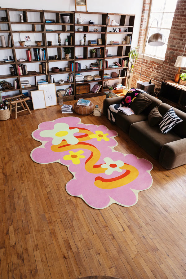 A playful rug with pink floral waves set on wood floor beside a dark sofa and tall shelves filled with open compartments.