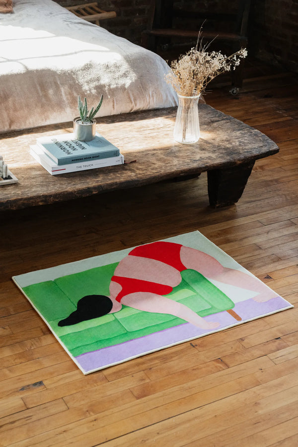 Modern abstract body rug in 2'X3', placed near a low wooden bench with dried flowers in a clear vase beside a cozy bed.