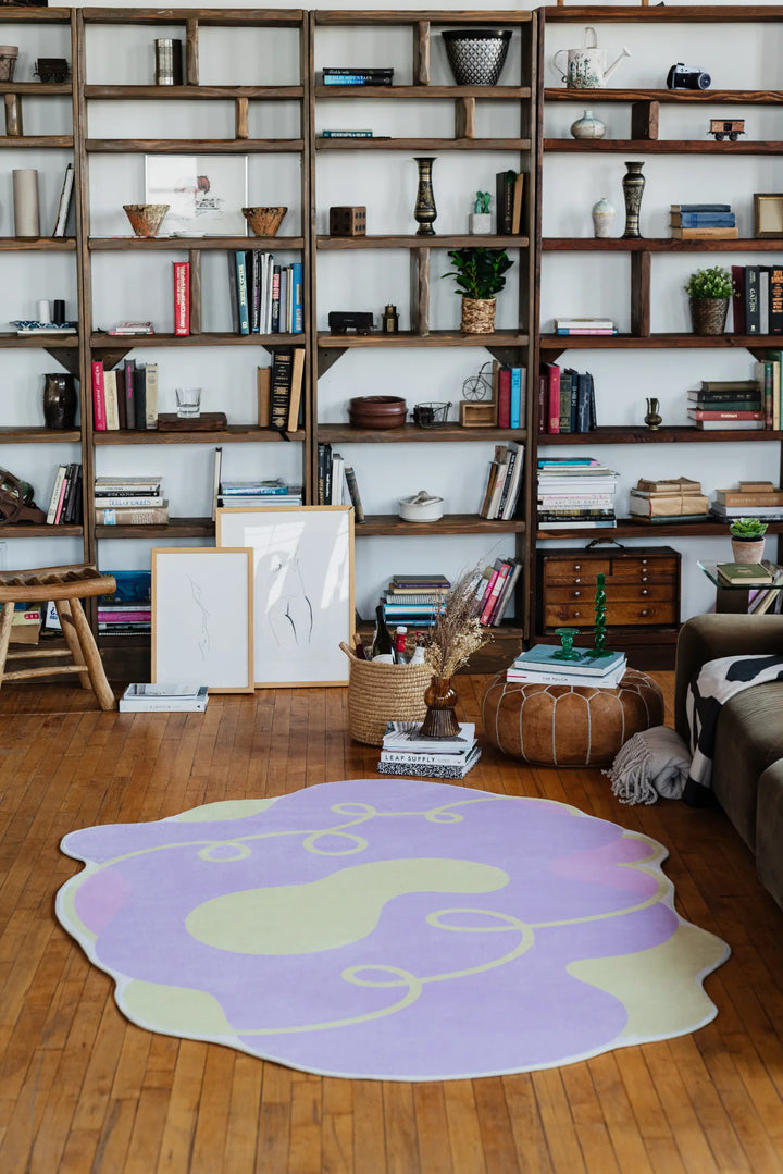 Lilac area rug with cream patches and looping yellow swirls, styled against a tall bookshelf and wooden floor for a cozy modern vibe.