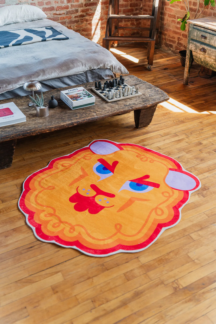 Cartoon lion head rug in bright orange and red tones placed on wood floor in front of a rustic wooden table.