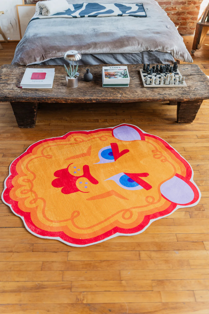 A playful lion rug with bold cartoon face resting on wood floor in front of a wooden bedside table and exposed brick wall.
