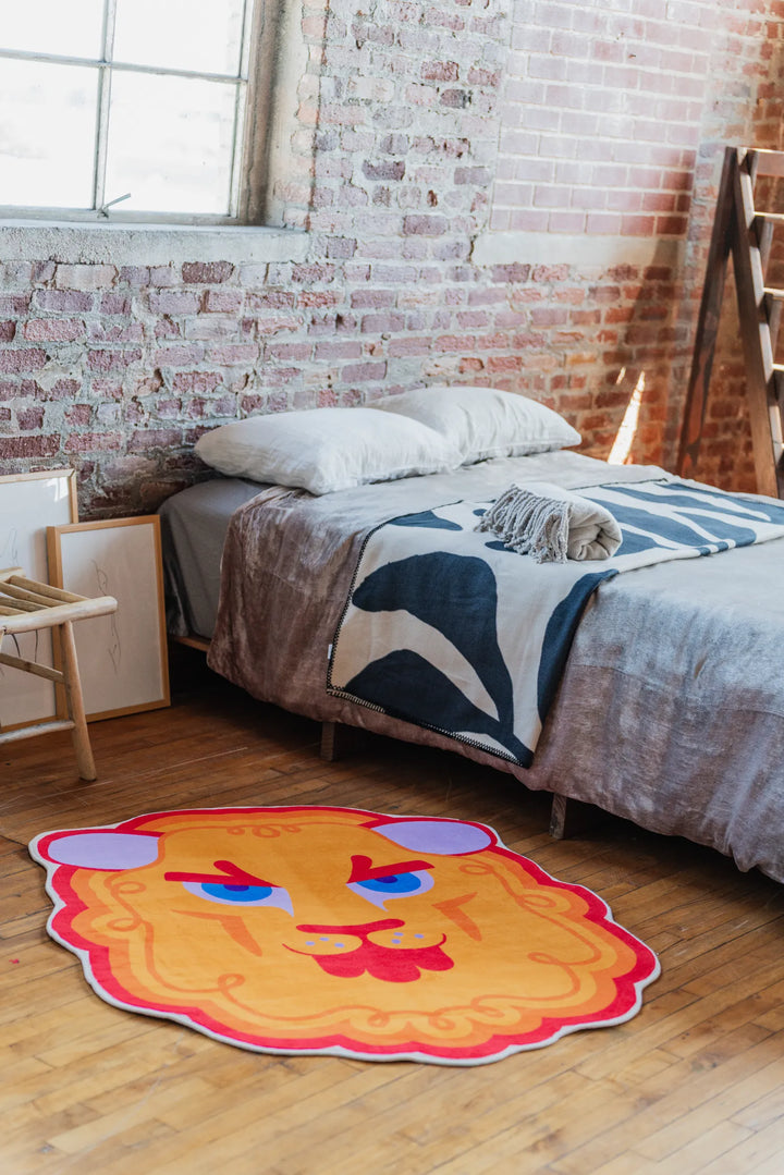 Cartoon lion head rug adds a bold accent on wood floor beside a simple bed and textured brick wall backdrop.
