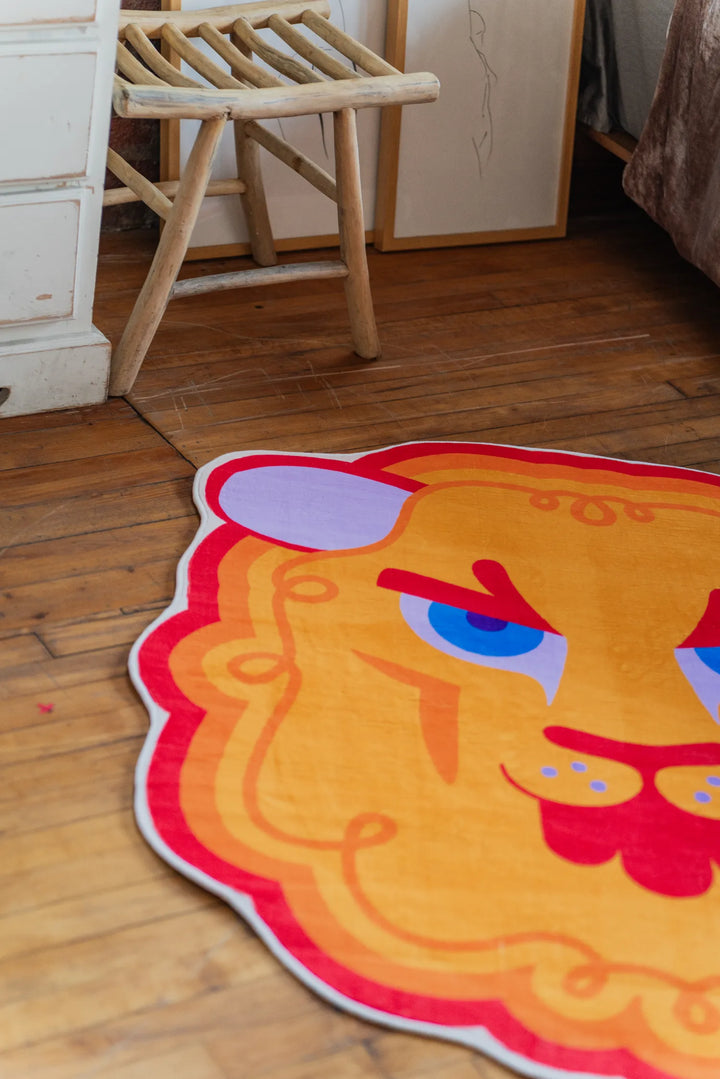 Close-up of cartoon lion head rug resting on wood floor beside a wooden chair and framed artwork leaning nearby.