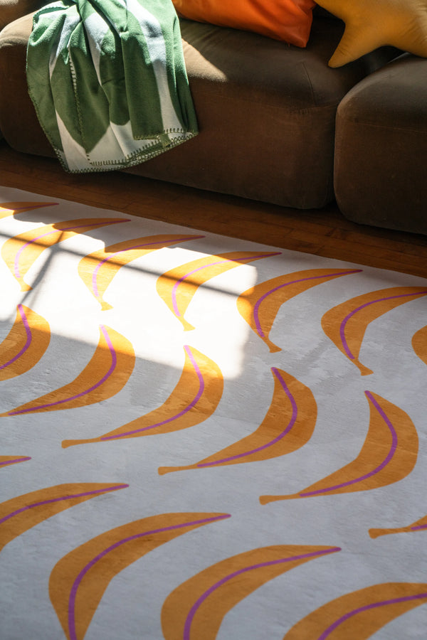 Close-up of banana-patterned funky rug catching sunlight beside a deep brown sofa on wooden flooring