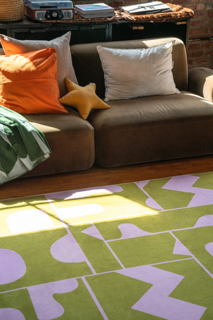 Close-up of olive green Abstract Patchwork Rug next to brown couch and throw pillows in modern apartment decor