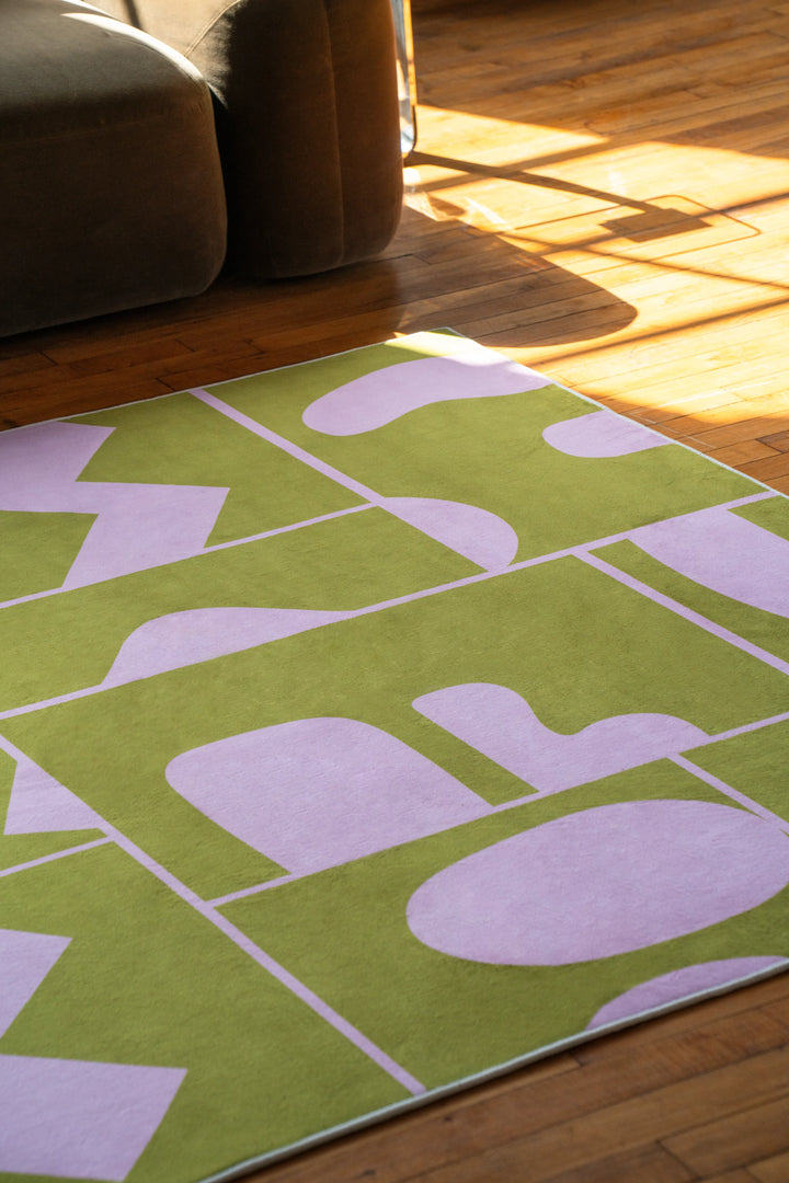 Corner view of olive green Abstract Patchwork Rug with soft purple shapes beside modern sectional seating