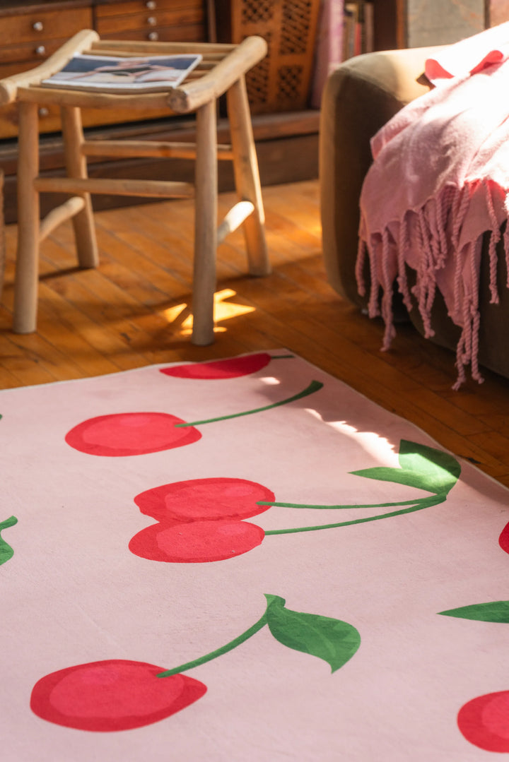 Cherry area rug in soft pink with vibrant red cherries and green stems, styled under a chair in a sunlit living space
