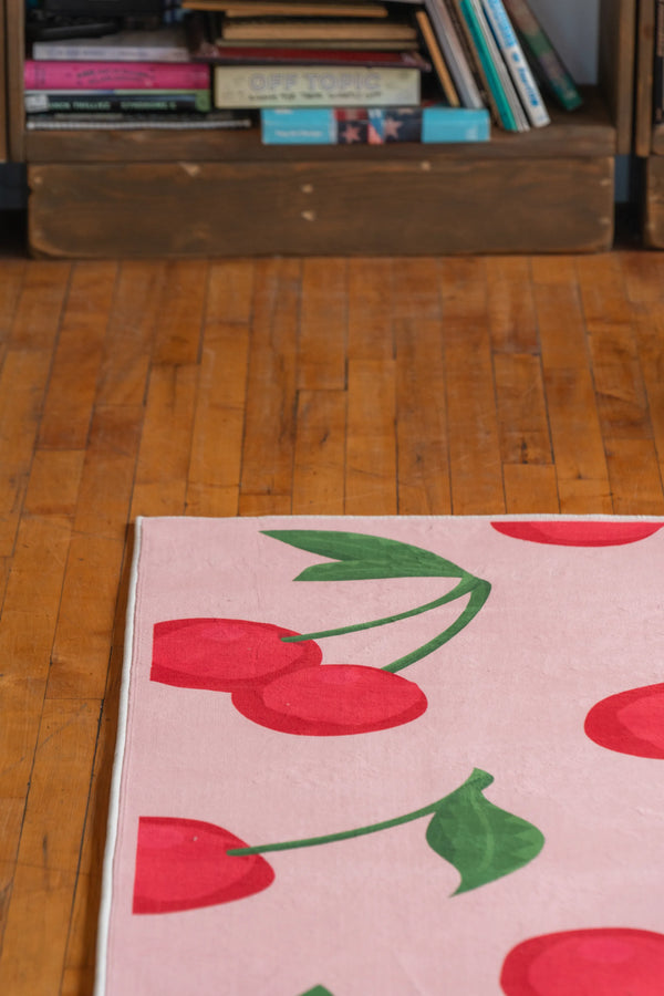 Corner detail of a cherry area rug with bright red fruit and bold green leaves on pastel pink background over wood floors