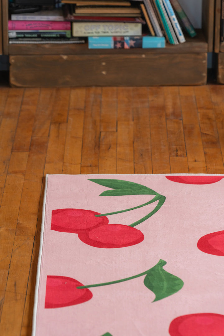 Corner detail of a cherry area rug with bright red fruit and bold green leaves on pastel pink background over wood floors