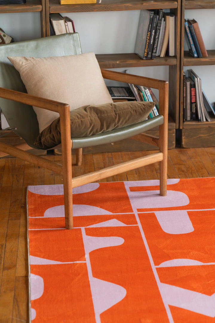 Abstract orange rug with geometric blocks placed below a minimalist lounge chair and shelf, bold modern rug design