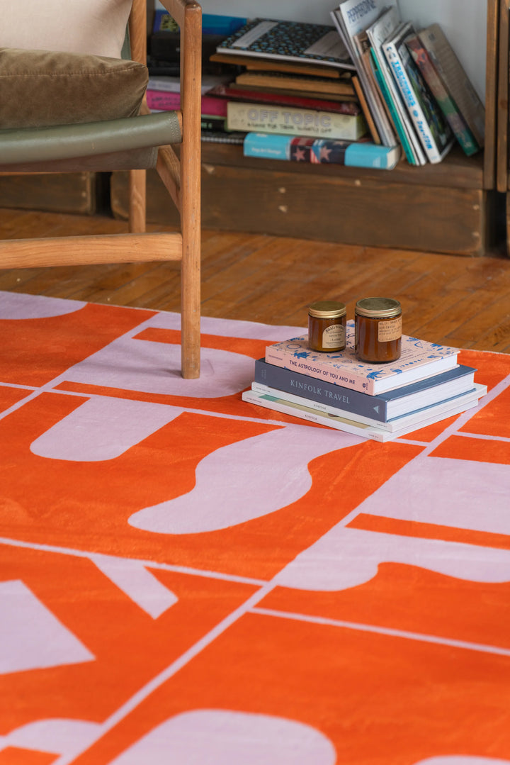Orange Geometric Patchwork Rug placed beneath a low green chair, beside a book-filled shelf on textured wood floor