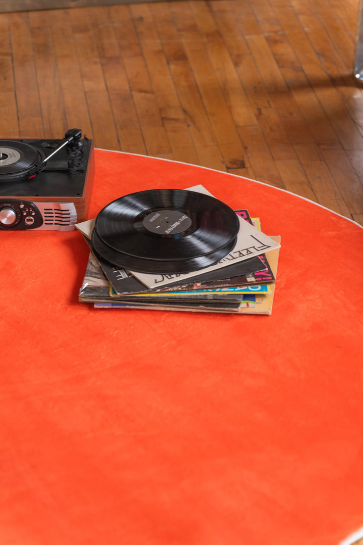 A stack of vinyl records and a turntable placed on the edge of an orange circular rug