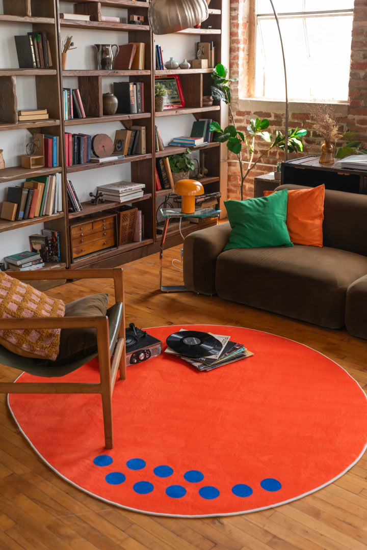 An orange circular rug with blue dots placed in a living room, under a turntable and books