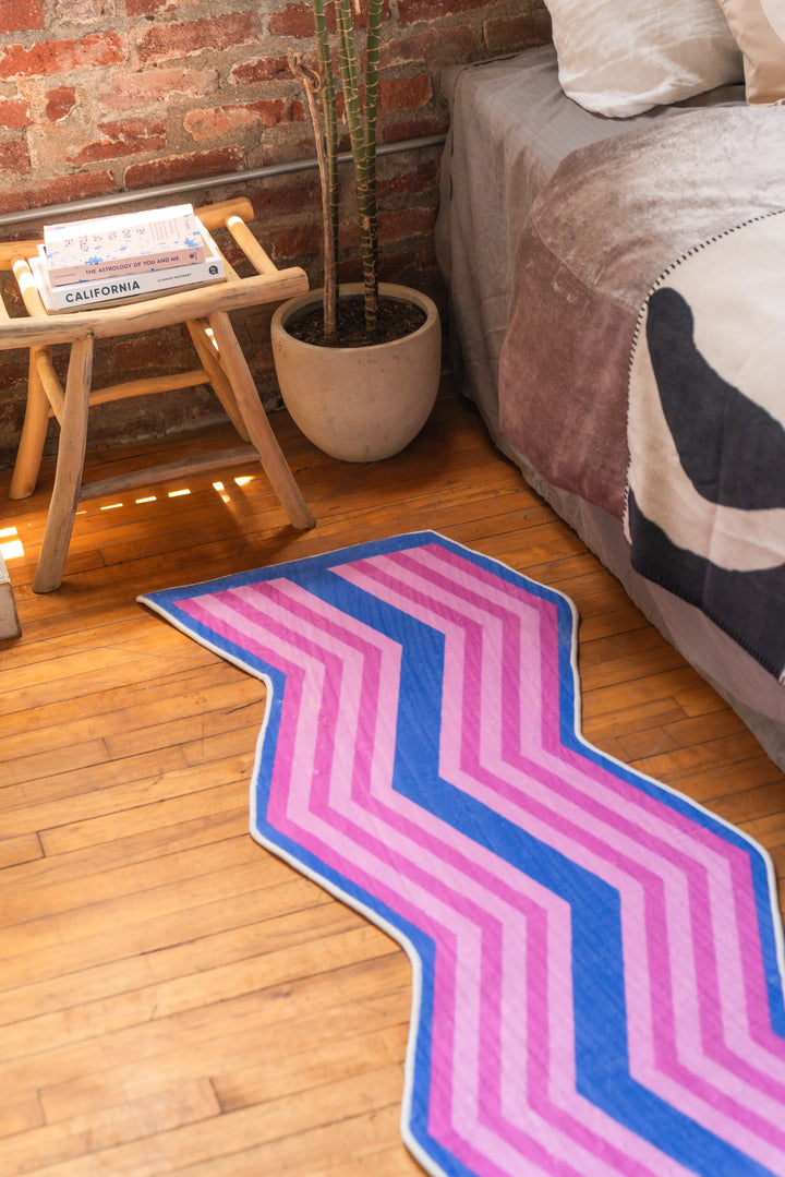 Wavy geometric area rug in pink and blue beside bed and wooden stool in brick-walled room