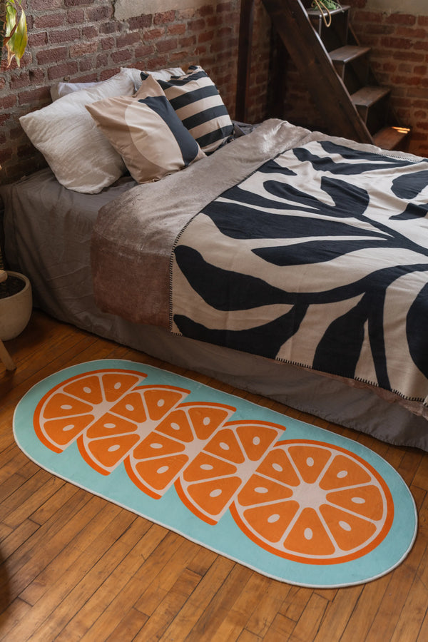 Oval orange slice rug on wooden floor beside bed with zebra-patterned bedding and beige throw, next to potted plant