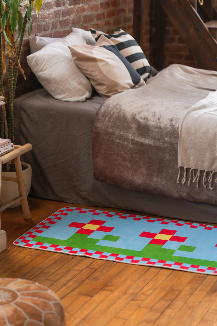 Multicolor pixel flower rug placed beside a bed with striped pillows and velvet bedding, on hardwood floor