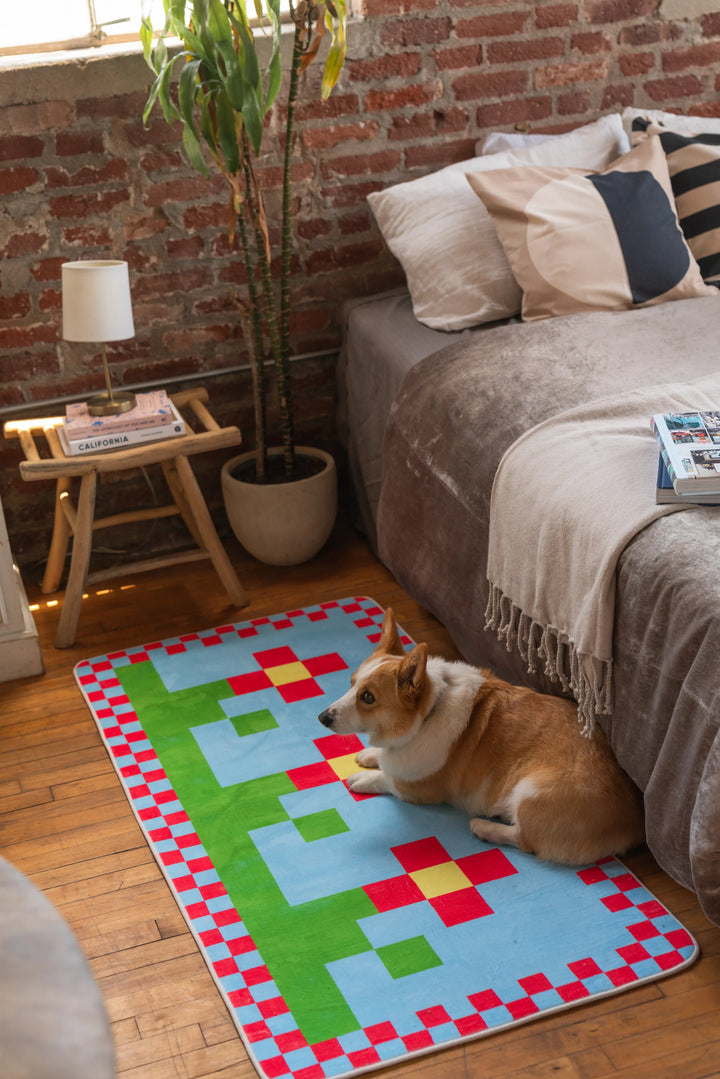 Corgi lying on a multicolor pixel flower rug next to a velvet-covered bed and round nightstand in a brick-wall bedroom