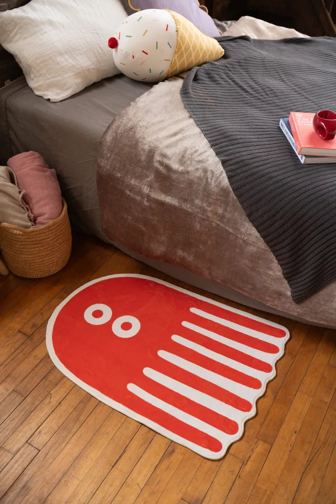 Small octopus-shaped rug with bold white stripes and eye motifs on orange, placed beside a bed with blanket and plush pillow