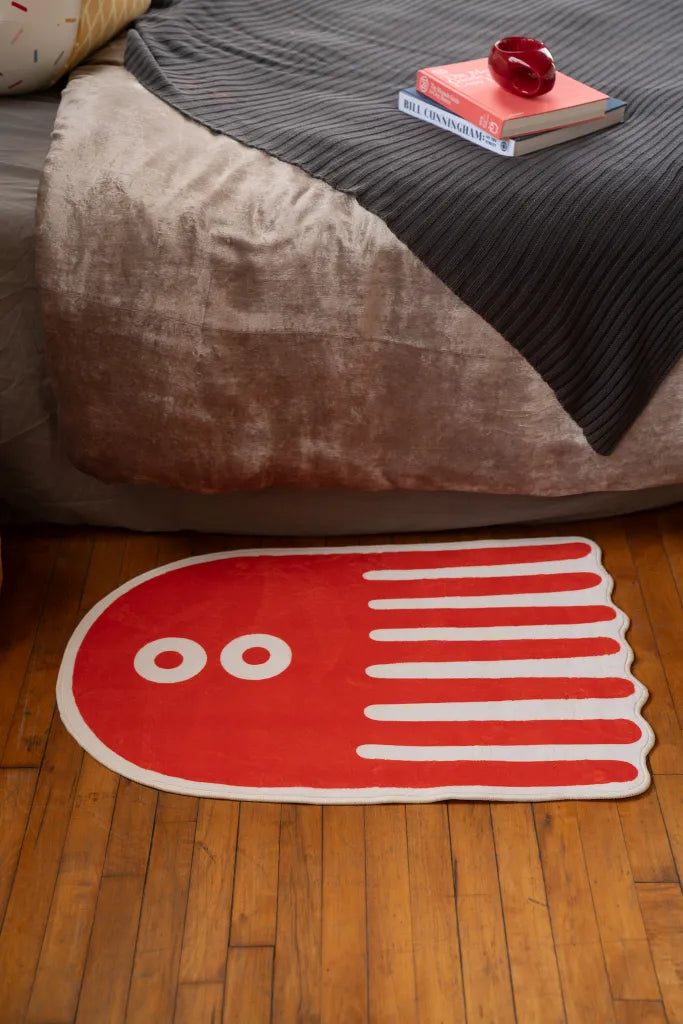 Playful orange rug with octopus-like shape and white stripe details, placed at the bedside on warm wooden floor