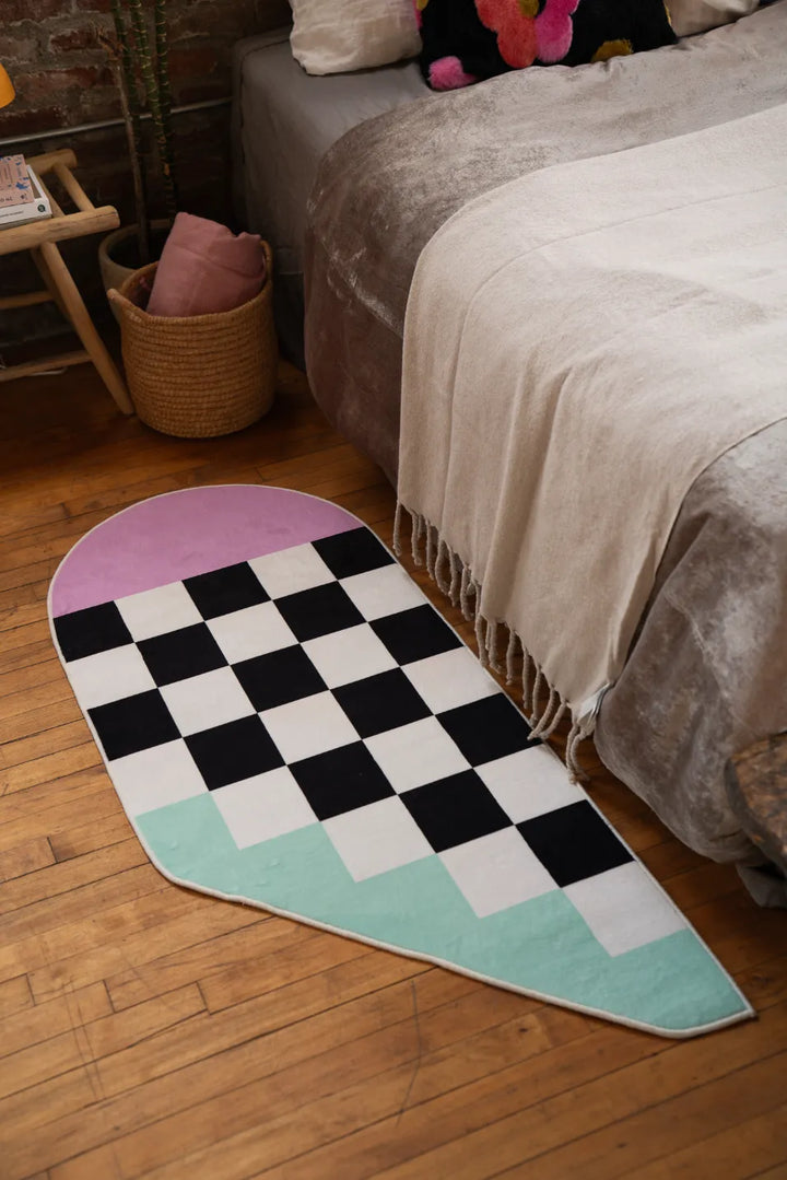 Asymmetrical checker rug set near the edge of a low bed on wood floor, softening the corner with playful contrast