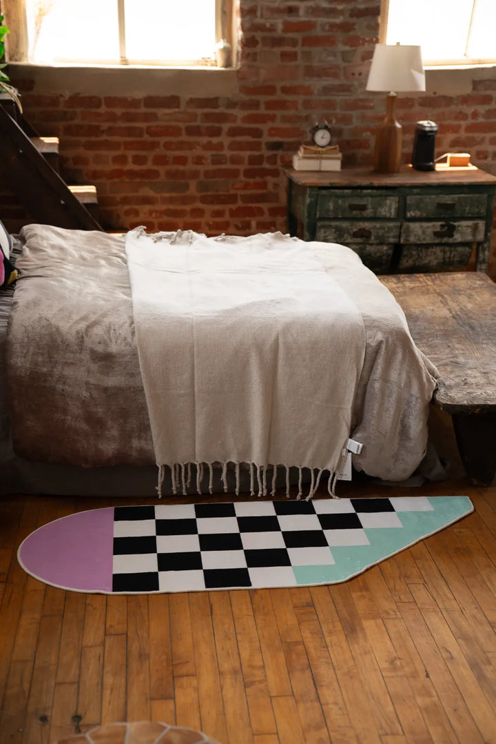 Pink and Blue Asymmetrical Checker Rug placed along the right side of a low bed, set on wood floor near an exposed brick wall