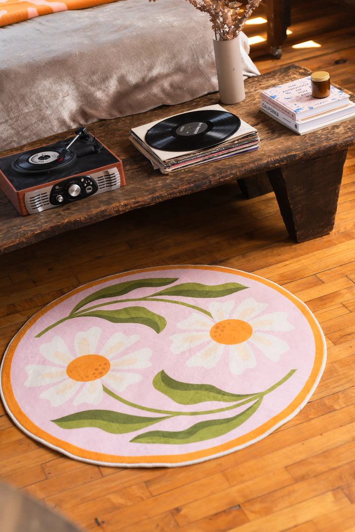 Dual daisy rug beside a distressed wood coffee table and soft upholstered seat, set on honey-toned wood floor.