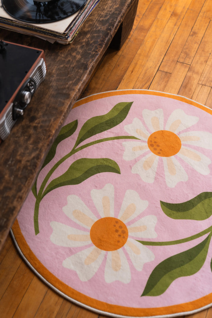 Close-up of daisy round rug with two white blooms and green stems beside a table corner on natural wood floor.